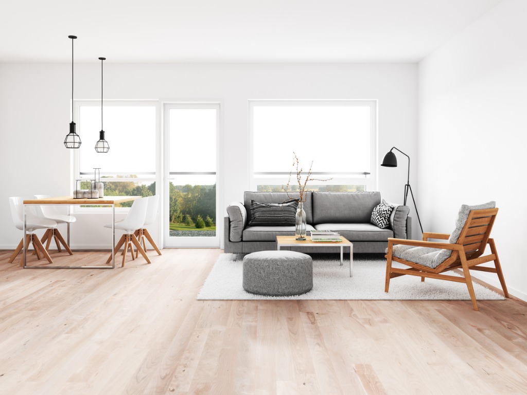 Open plan room with light wood floors showing living area with grey sofa, wood armchair, grey ottoman on rug, black floor lamp, and dining area with wood/white table, white chairs, and two black pendant lights; large windows overlook trees. Open plan room with light wood floors showing living area with grey sofa, wood armchair, grey ottoman on rug, black floor lamp, and dining area with wood/white table, white chairs, and two black pendant lights; large windows overlook trees.