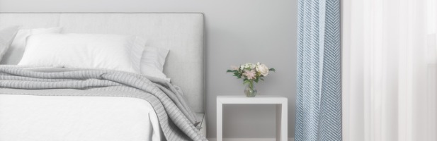 Interior view of a modern bedroom showcasing a grey upholstered headboard, crisp white bedding with a grey knit throw, a white nightstand holding a small floral arrangement, and light blue curtains with a subtle pattern. Interior view of a modern bedroom showcasing a grey upholstered headboard, crisp white bedding with a grey knit throw, a white nightstand holding a small floral arrangement, and light blue curtains with a subtle pattern.