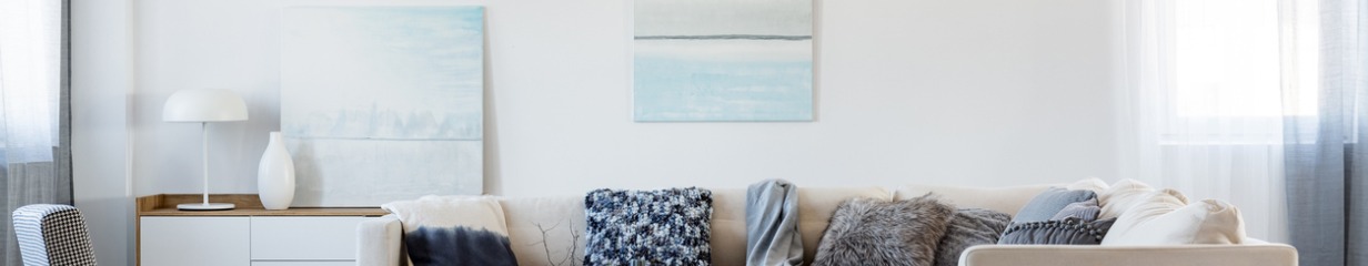 Close-up of a modern living room showing a cream sofa adorned with diverse pillows (patterned blue, dip-dye, faux fur grey), beside a white sideboard with a lamp and vase, under two abstract blue and white paintings. Close-up of a modern living room showing a cream sofa adorned with diverse pillows (patterned blue, dip-dye, faux fur grey), beside a white sideboard with a lamp and vase, under two abstract blue and white paintings.