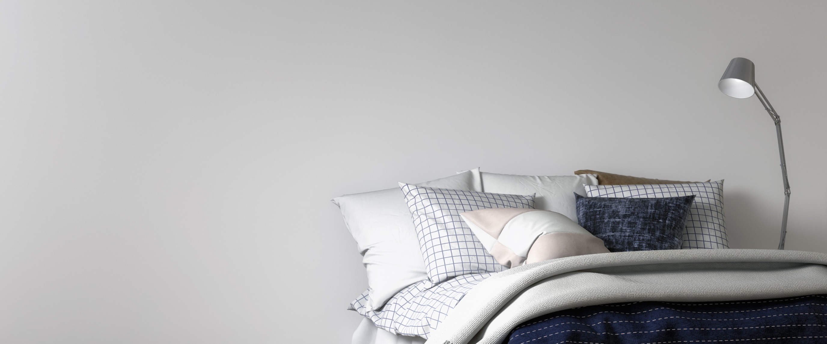 Close-up of bed head: pillows stacked include white, blue/white check, dark blue textured, and pink geometric; bedding features folded dark blue coverlet over grey sheet; a modern adjustable metal floor lamp stands beside.