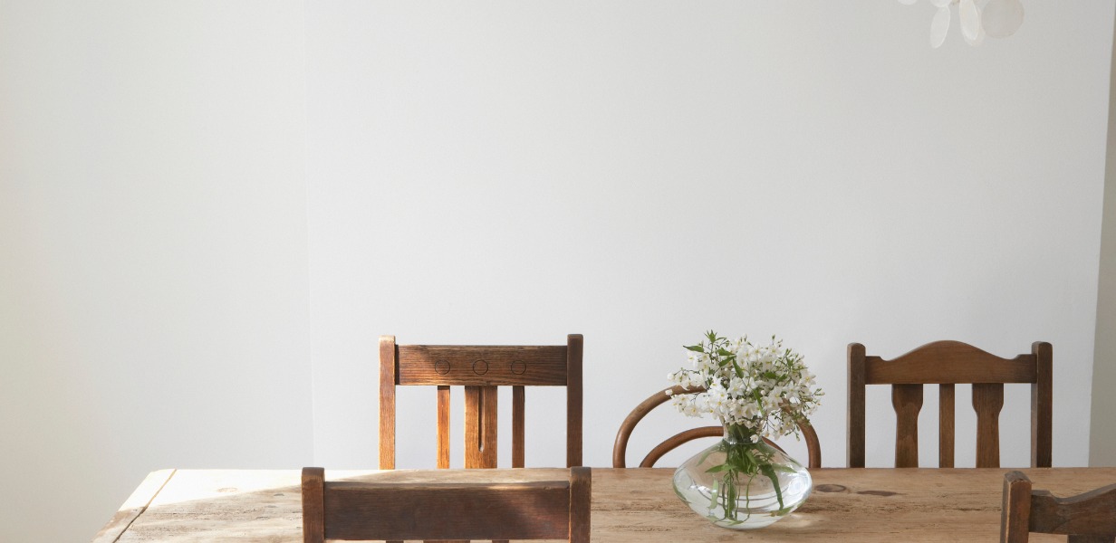 Close-up of a sunlit rustic wooden dining table showing wood grain, with dark wood chairs featuring vertical back slats; a round glass vase with small white flowers sits on the table.