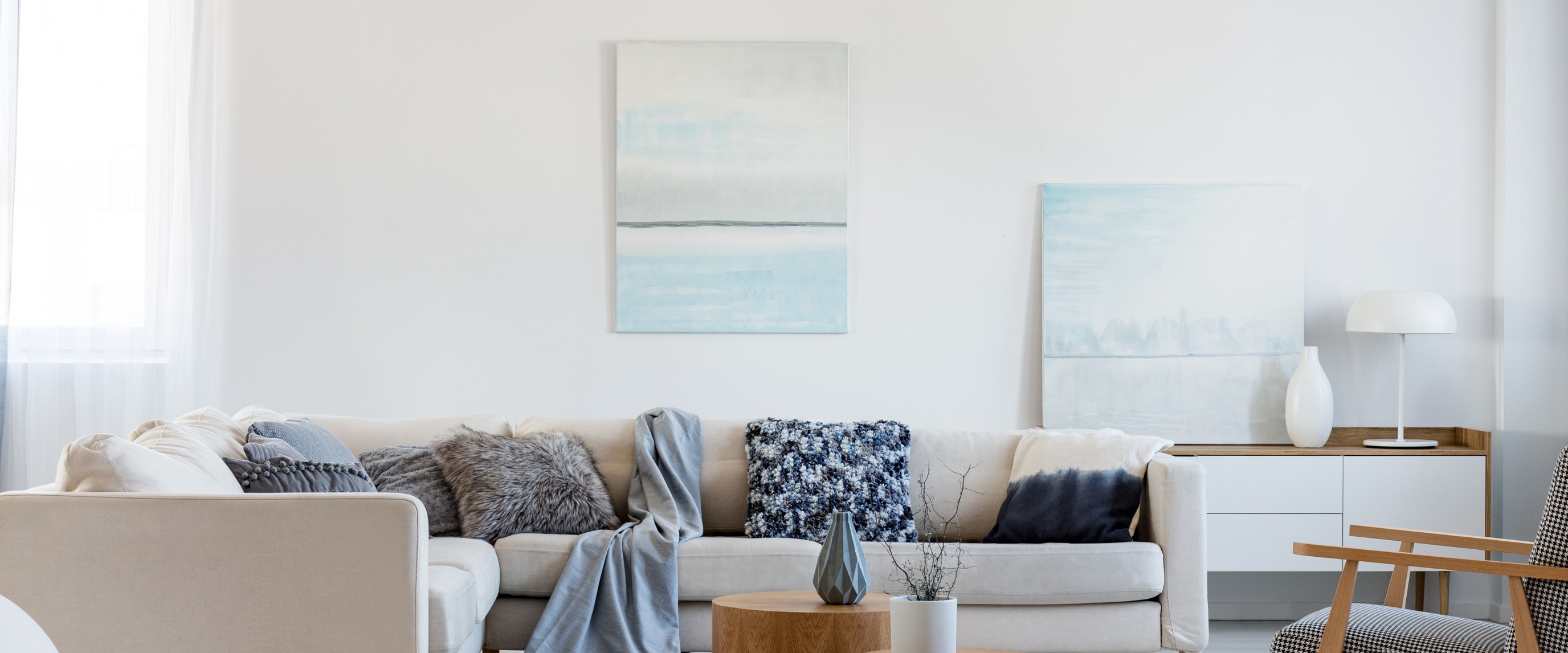 Living room with cream L-shaped sectional sofa displaying diverse pillows (patterned blue, dip-dye, furry grey), round wood coffee table with vase, white sideboard with lamp on the right, and two abstract blue/white paintings.